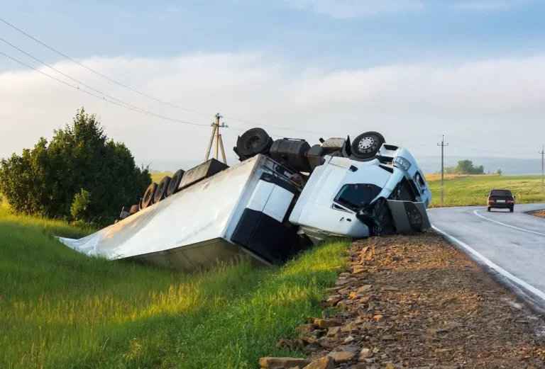 An overturned white truck lying on its side in a grassy roadside ditch with a car passing on the road, blue sky and green grass, calm rural mood