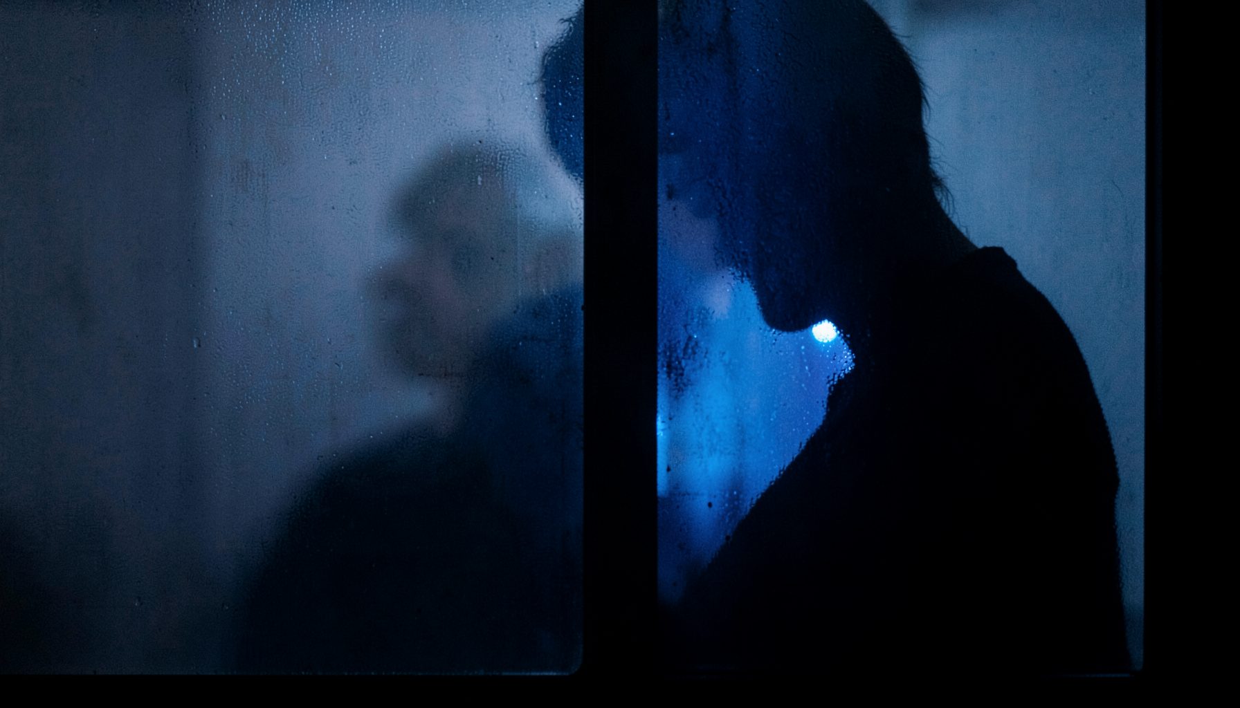 A silhouette of a person peering through a frosted window pane at night, with faint blue light spilling through.