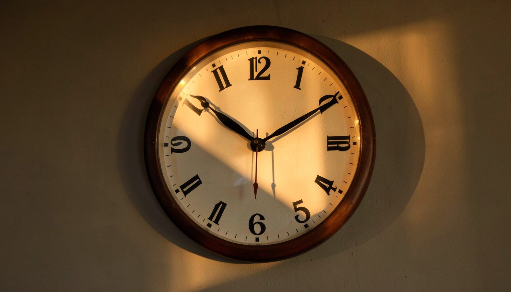 A wall clock showing an evening hour, with soft golden-hour light streaming across the face, casting long shadows.