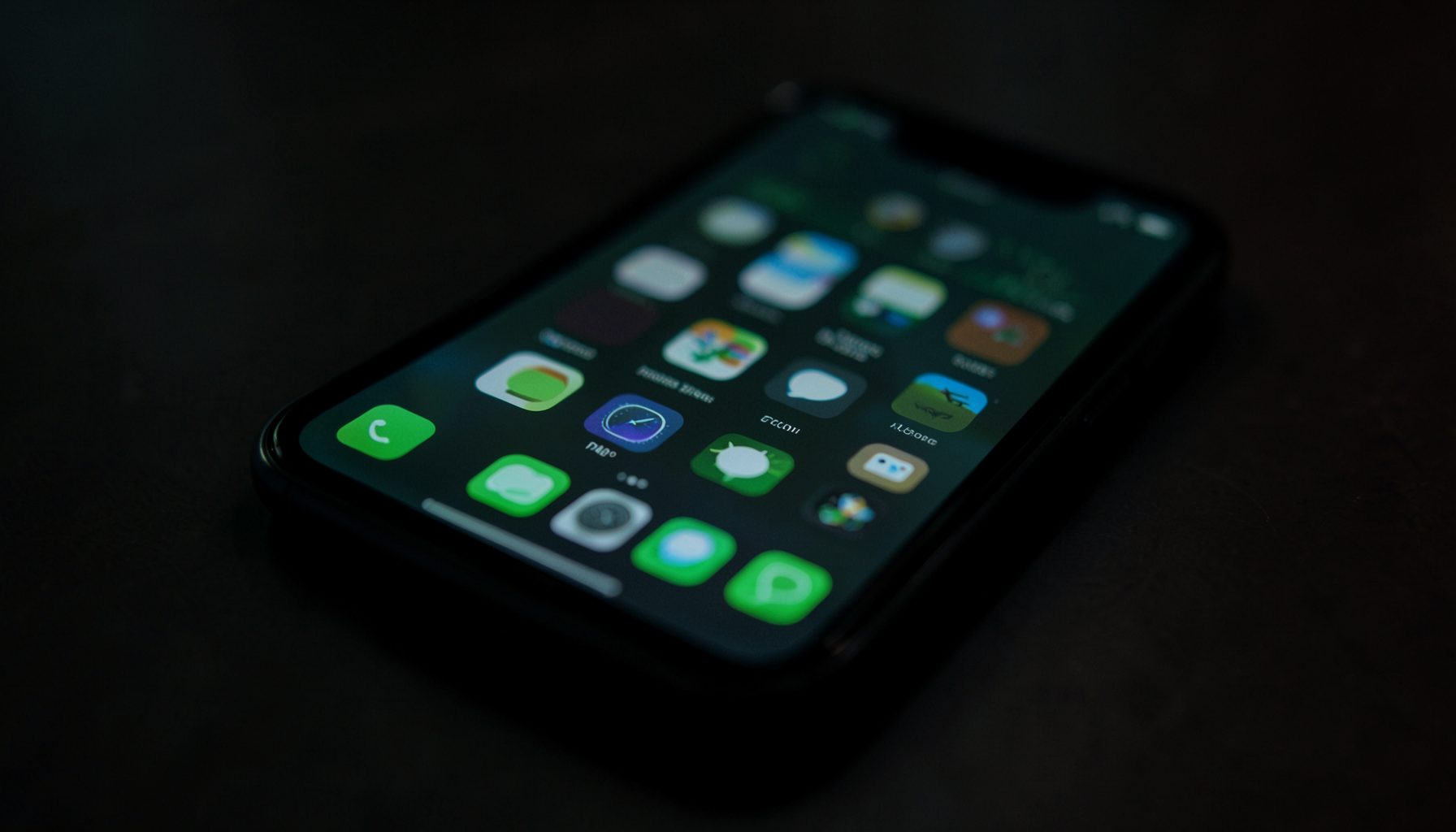 A dimly lit smartphone screen displaying a grid of app icons in green and dark tones, resting on a dark surface with soft shadows