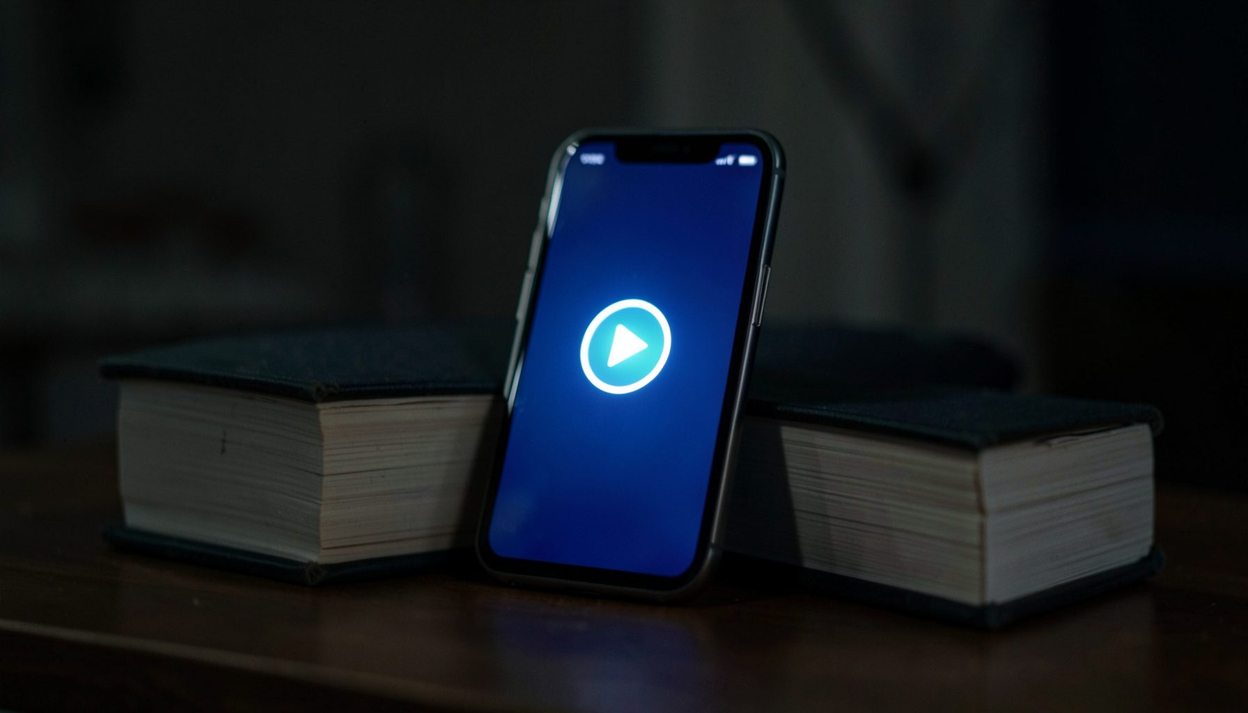 A smartphone propped against a stack of books, its screen glowing with a video play button icon in a dimly lit room.